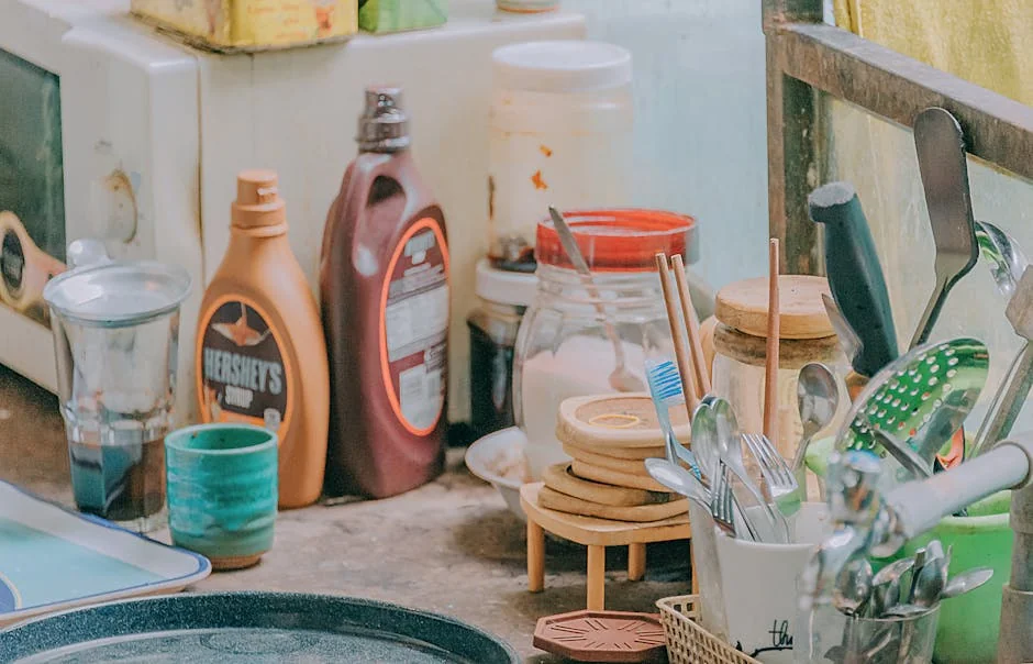 Close-up of a cluttered kitchen counter with utensils, jars, and a Hershey's syrup bottle. - best budgetfriendly daily essentials for homeowners 2026