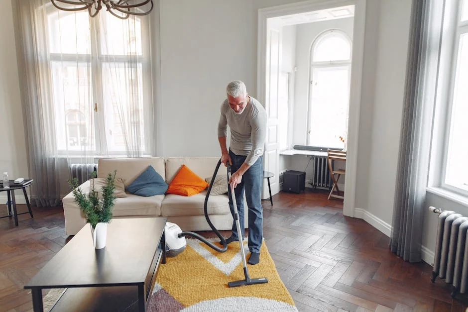 From above of focused grey haired man with beard vacuum cleaning carpet at home - best daily essentials for elderlyfriendly home living