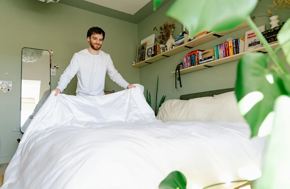 Man makes his bed in a bright, green room. - what to look for when purchasing highquality bed linens